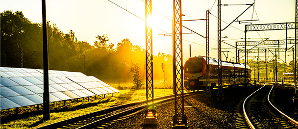 Solar panels beside the ŁKA railway line at sunset, with a train passing under the catenary